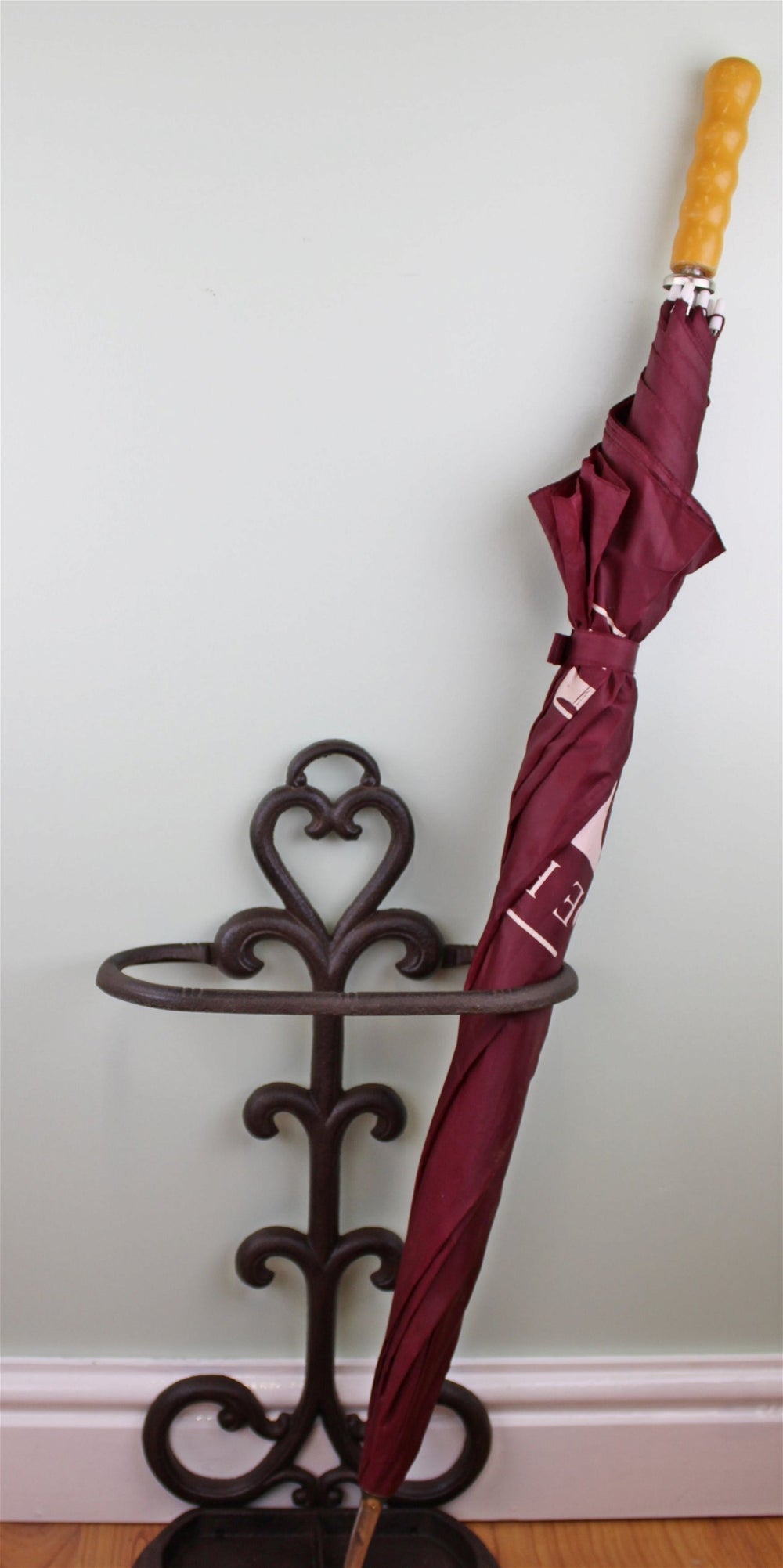 A maroon umbrella with a light brown handle is neatly stored in a Traditional Rustic Cast Iron Umbrella Stand featuring a heart-shaped design, placed against a light-colored wall near a wooden floor, ensuring a drip-free hallway.