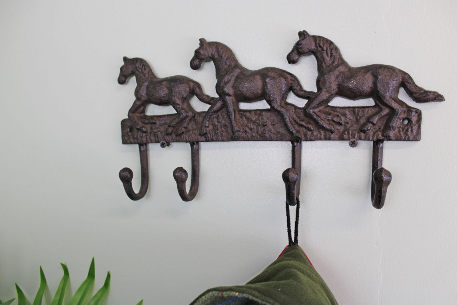 The Rustic Cast Iron Wall Hooks, Three Horses, is a dark metal wall-mounted coat rack that showcases a decorative design featuring three running horses and includes three hooks. Ideal for small space storage accessories, it is mounted on a plain white wall with some green leaves visible in the bottom left corner.