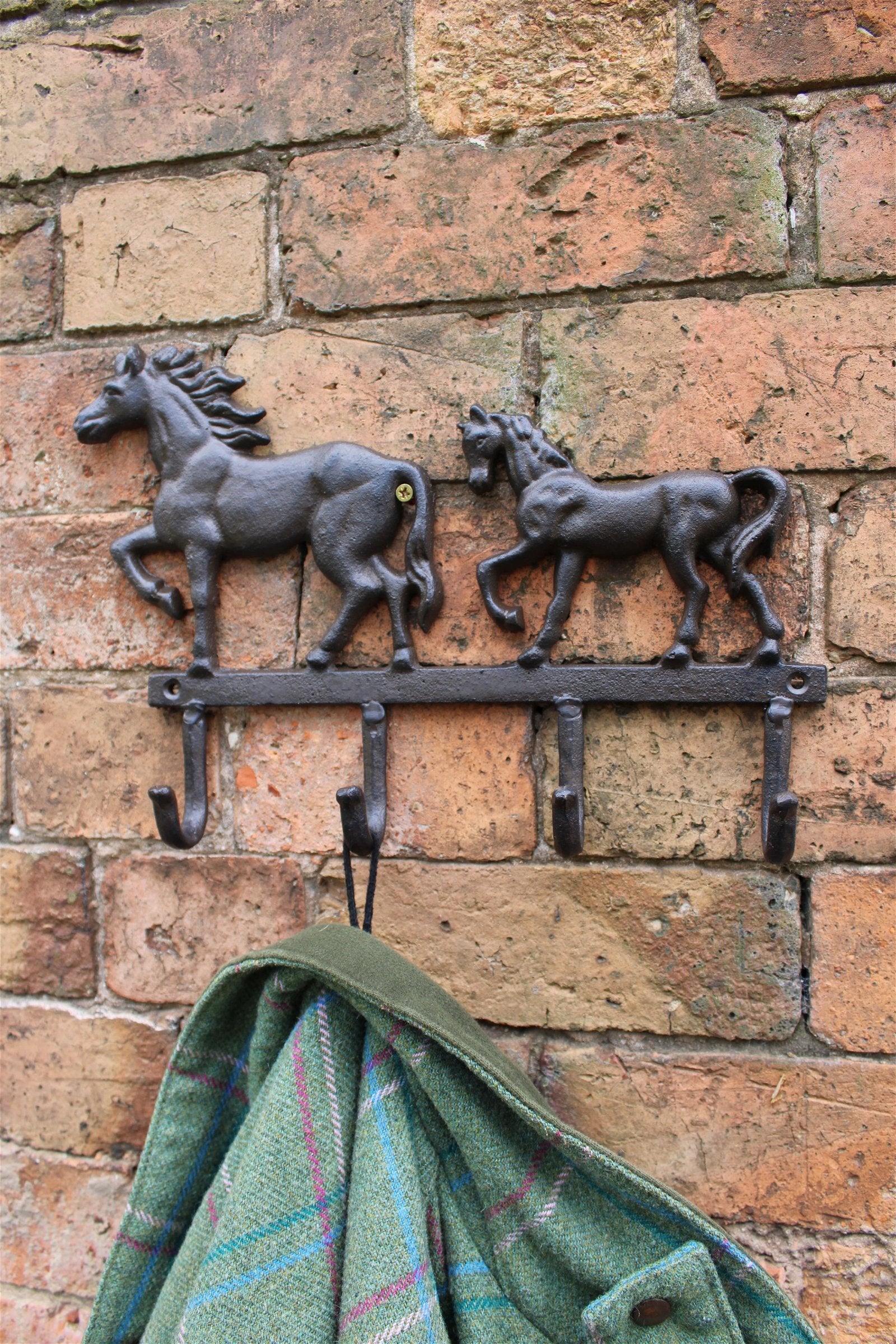 The Rustic Cast Iron Wall Hooks with Two Horses are mounted on a brick wall, showcasing two horse figurines that exemplify rustic charm. Below the horses, there are four hooks, one of which holds a green and plaid garment. The horses and hooks are made from the same dark cast iron material, enhancing the industrial style decor with functional elegance.