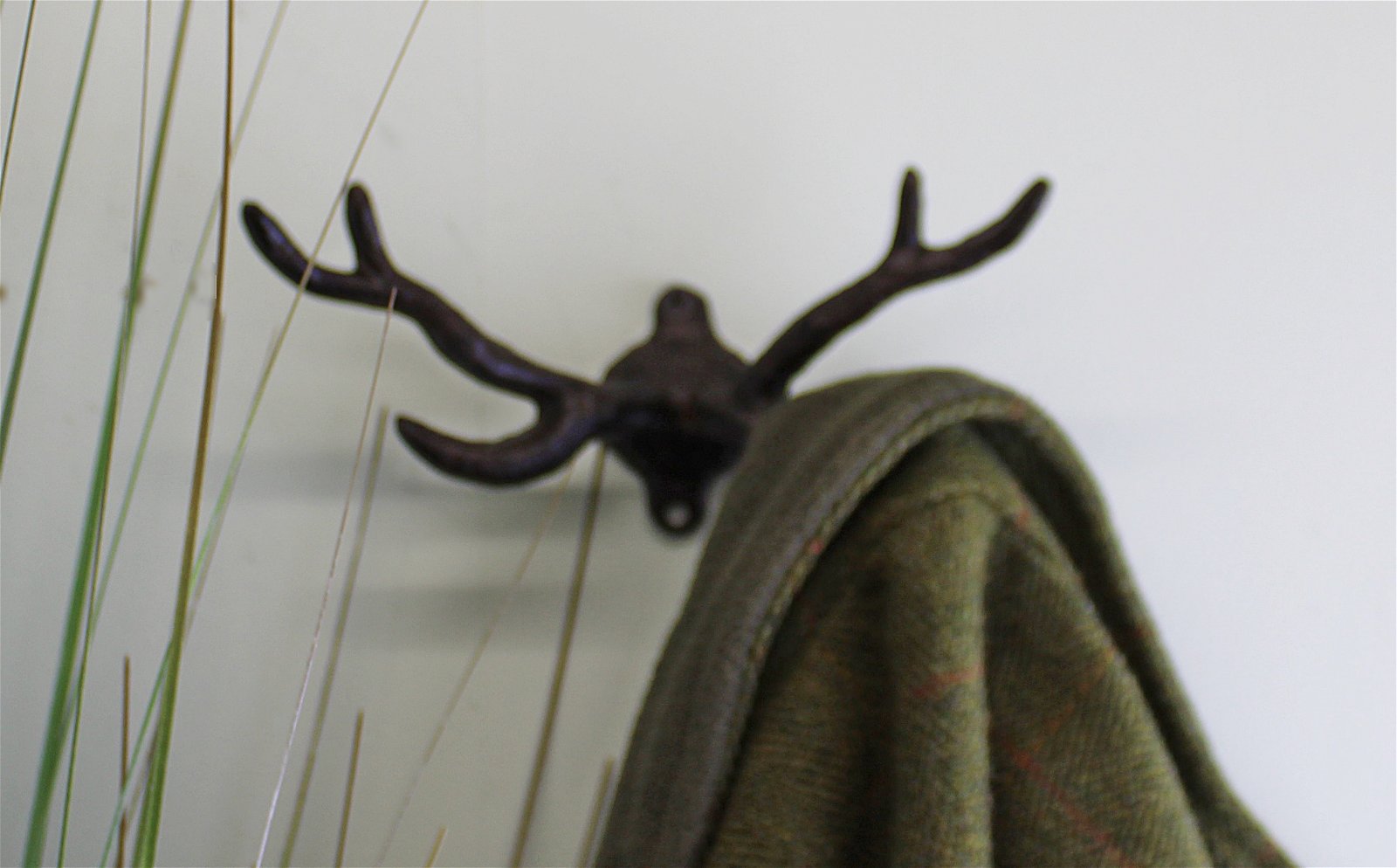 A wall-mounted Rustic Cast Iron Wall Hook in the shape of Stag Antlers is holding a green fabric, possibly a blanket or coat. Thin branches or decorative reeds are visible to the left of the hook, adding a natural, rustic touch to the industrial-style decor. Perfect for small space storage accessories.