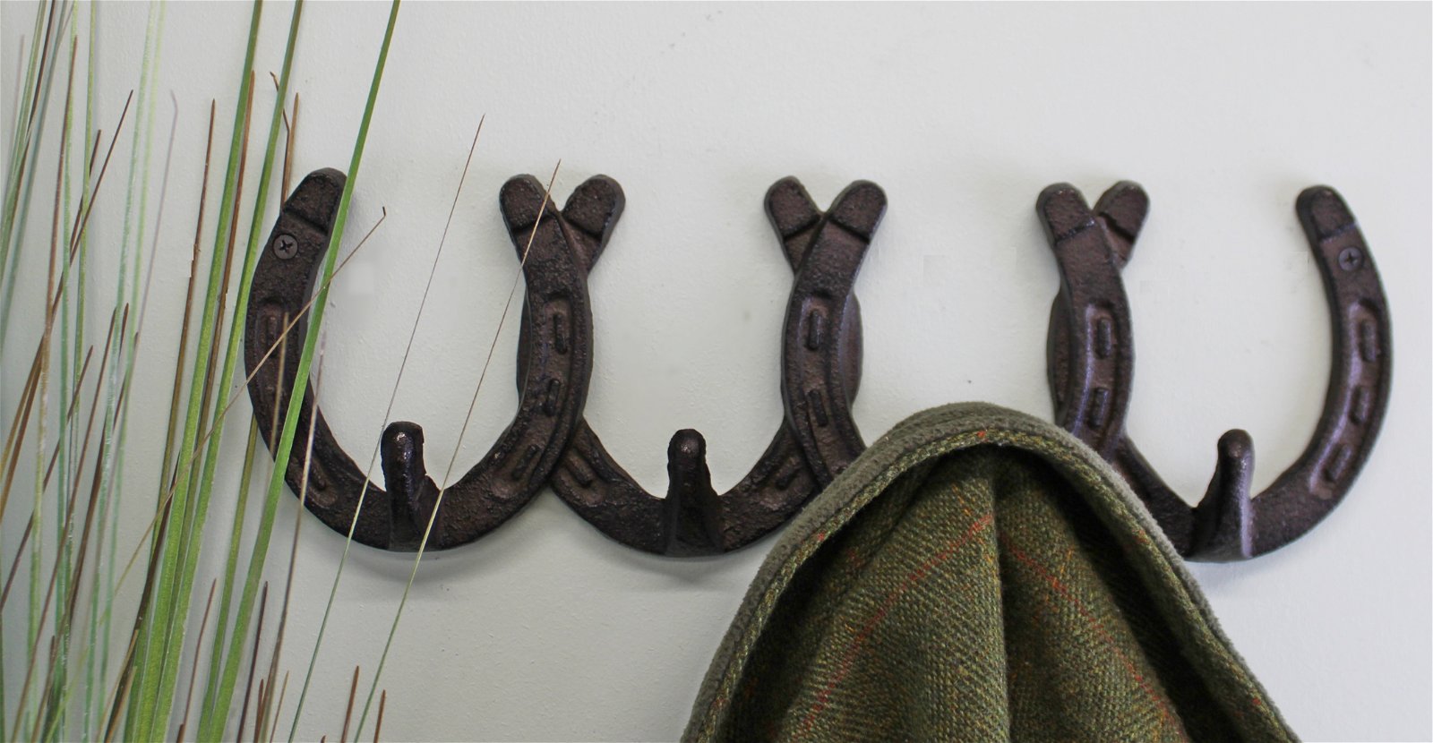 A coat rack featuring the Rustic Cast Iron Wall Hooks, Horseshoe Design, is mounted on a wall with a green and brown plaid jacket hung on one of the hooks. Tall grass stalks are placed to the left side, enhancing this industrial-style wall decor with a touch of nature.