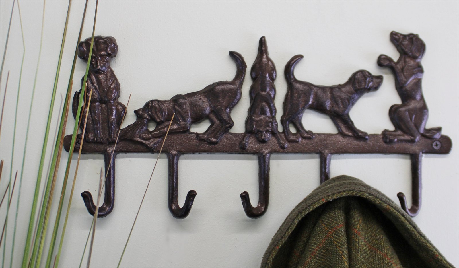 A decorative Rustic Cast Iron Wall Hook featuring five playful dog figures in various poses. Some dogs are standing, sitting, or sniffing the ground. The hooks below them hold a green plaid garment. Tall plant stems are visible on the left side, adding charm to your small space storage accessories.