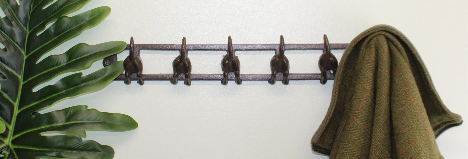 A rack featuring six rustic cast iron wall hooks shaped like dog tails sits mounted on a wall next to a large green tropical leaf. A greenish-brown cloth is hung over the hook on the far right, with the rest of the hooks unoccupied, offering industrial-style small space storage.