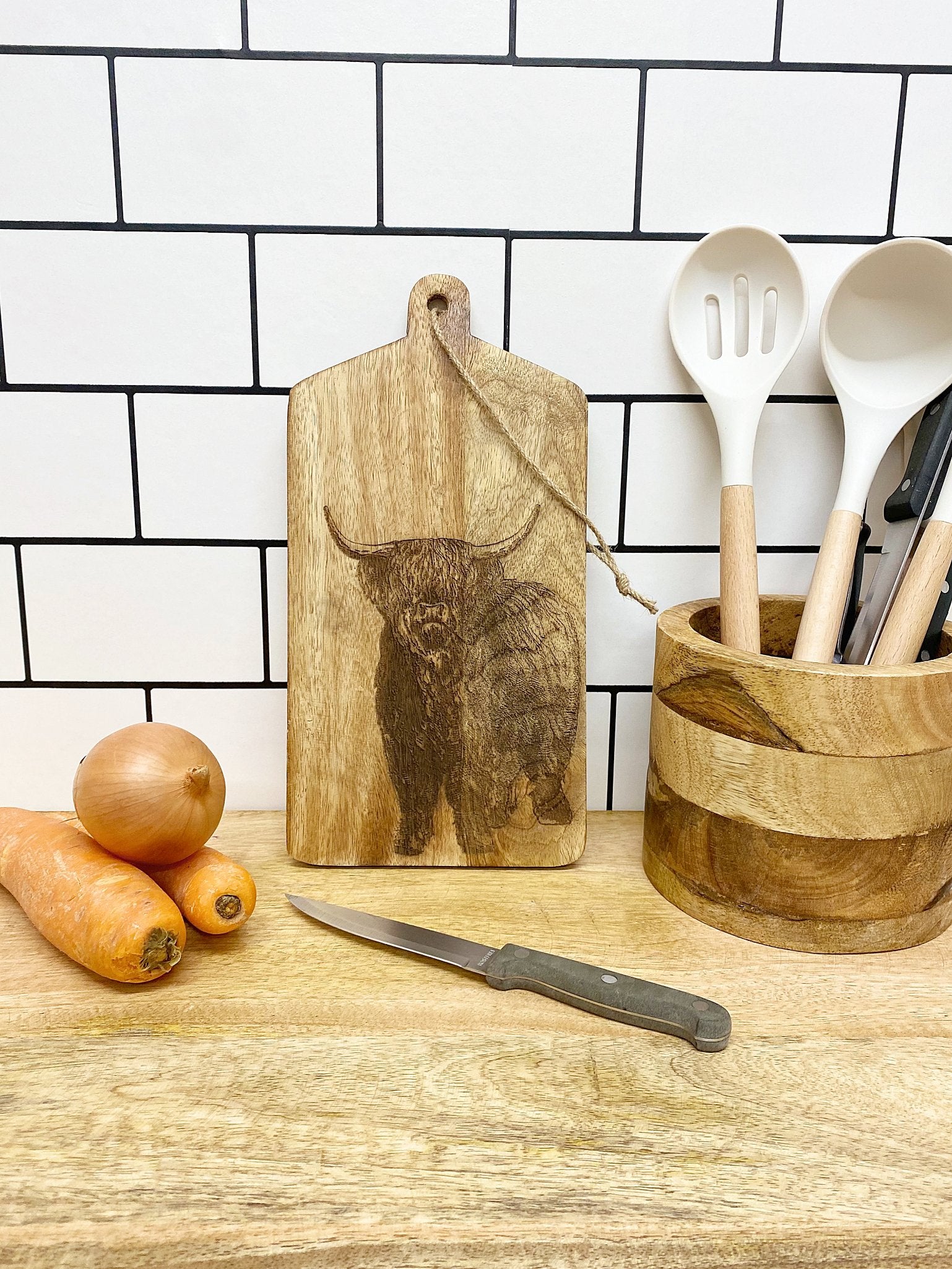 A Highland Cow Engraved Wooden Cheese Board showcasing a detailed bull illustration, accompanied by a knife, carrot, and onion. To the right, utensils such as spoons and a ladle are neatly organized in an engraved portrait wooden holder. The backdrop features a white tiled wall with black grout lines.