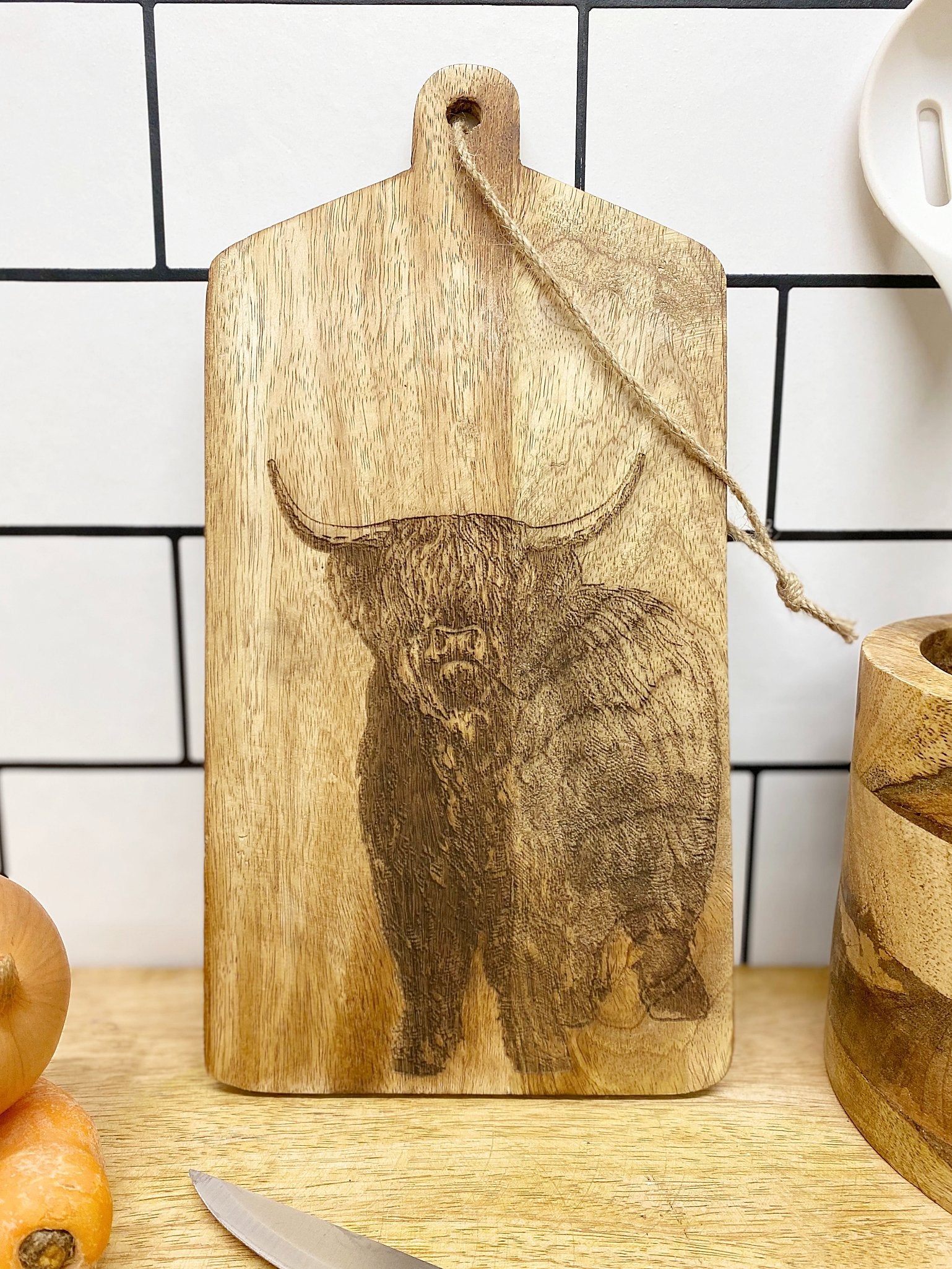 A Highland Cow Engraved Wooden Cheese Board rests against a tiled white backsplash. Surrounding the wooden cheese board are kitchen items, including a knife, an onion, a carrot, and a wooden utensil holder.