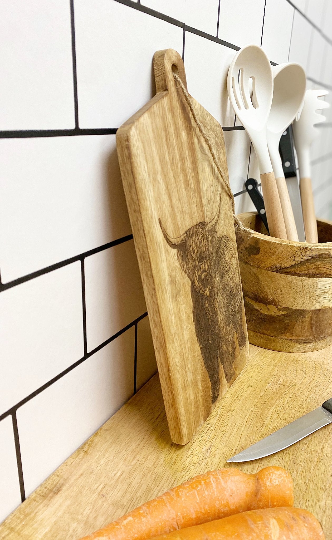 A kitchen scene showcases the Highland Cow Engraved Wooden Cheese Board, beautifully crafted from mango wood and featuring a detailed portrait of a highland cow. Nearby, white utensils rest in a wooden holder, and a few peeled carrots lie on the counter next to a knife. The backdrop is set with white subway tiles accented with black grout.