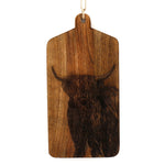 The Highland Cow Engraved Wooden Cheese Board is a rectangular mango wood cutting board with a handle, showcasing an intricately engraved portrait of a Highland cow with long horns and shaggy fur. The board can be conveniently hung by the loop of twine attached to the handle.
