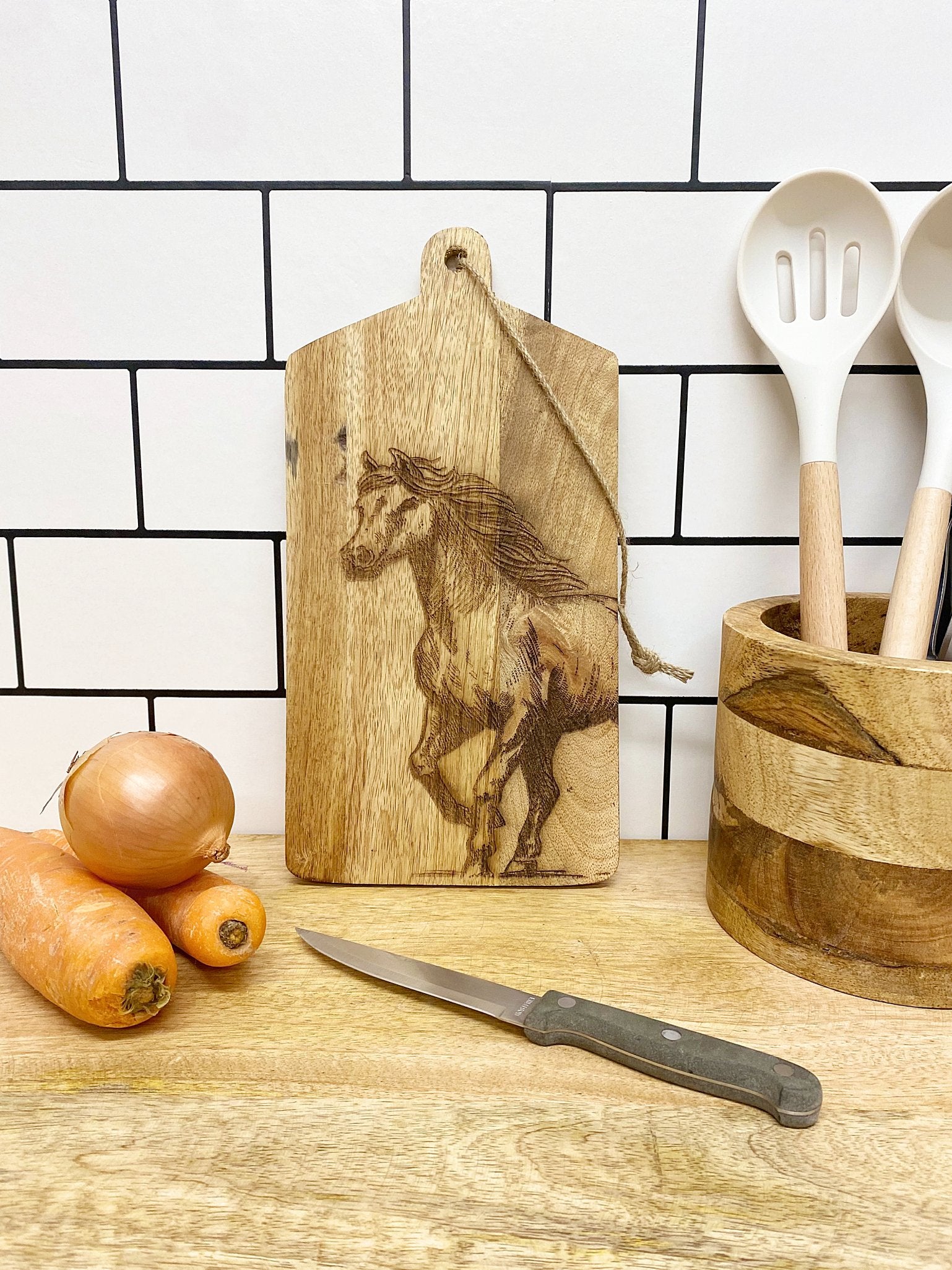 In a wooden kitchen scene, a Horse Engraved Wooden Cheese Board displays two carrots and an onion on the counter. A knife lies nearby, while a utensil holder with spoons is set against the white tile backsplash.