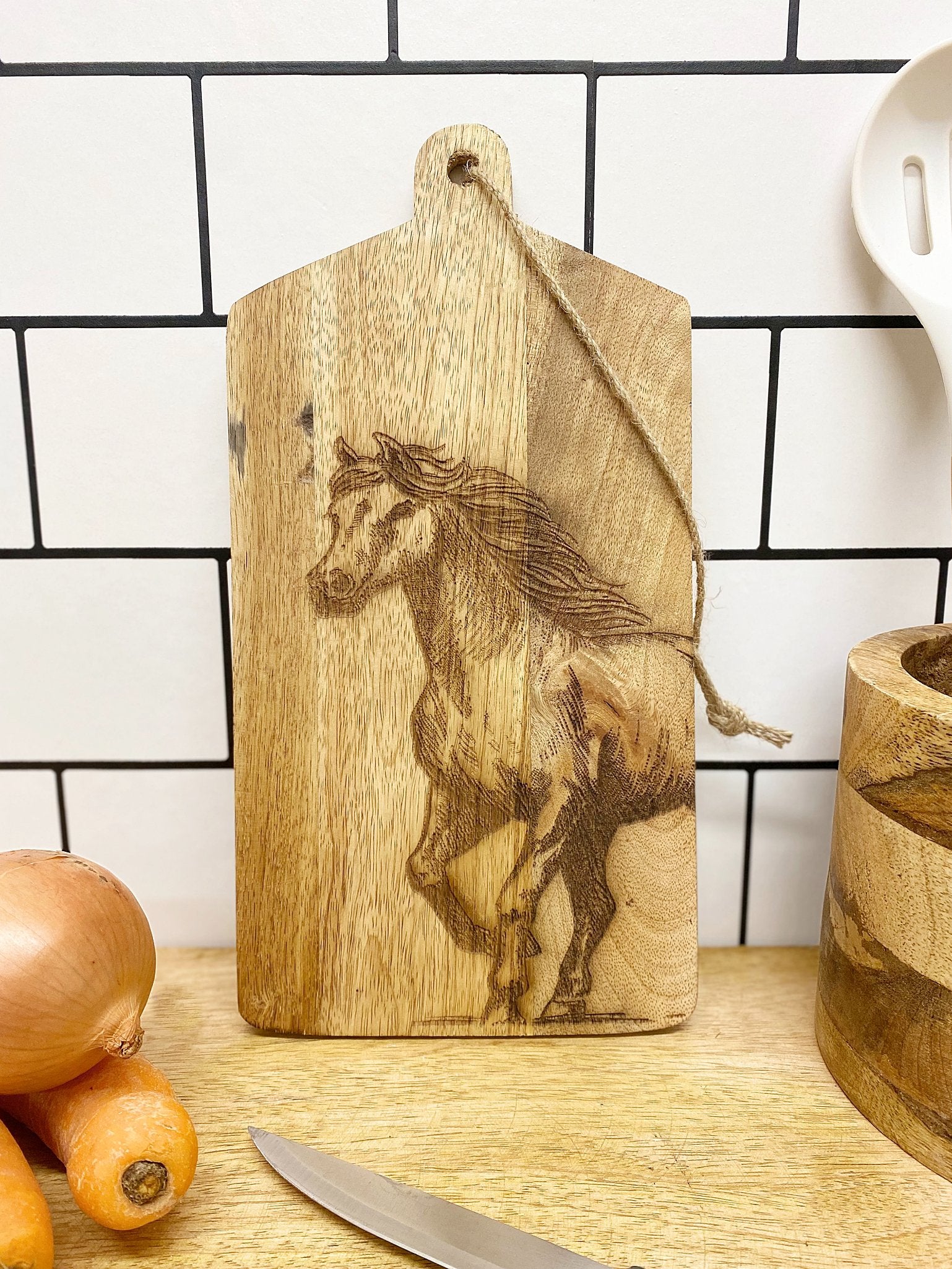 The Horse Engraved Wooden Cheese Board, made from mango wood, hangs by a string against the white tile background of the kitchen counter, surrounded by onions, carrots, a knife, and a wooden bowl.