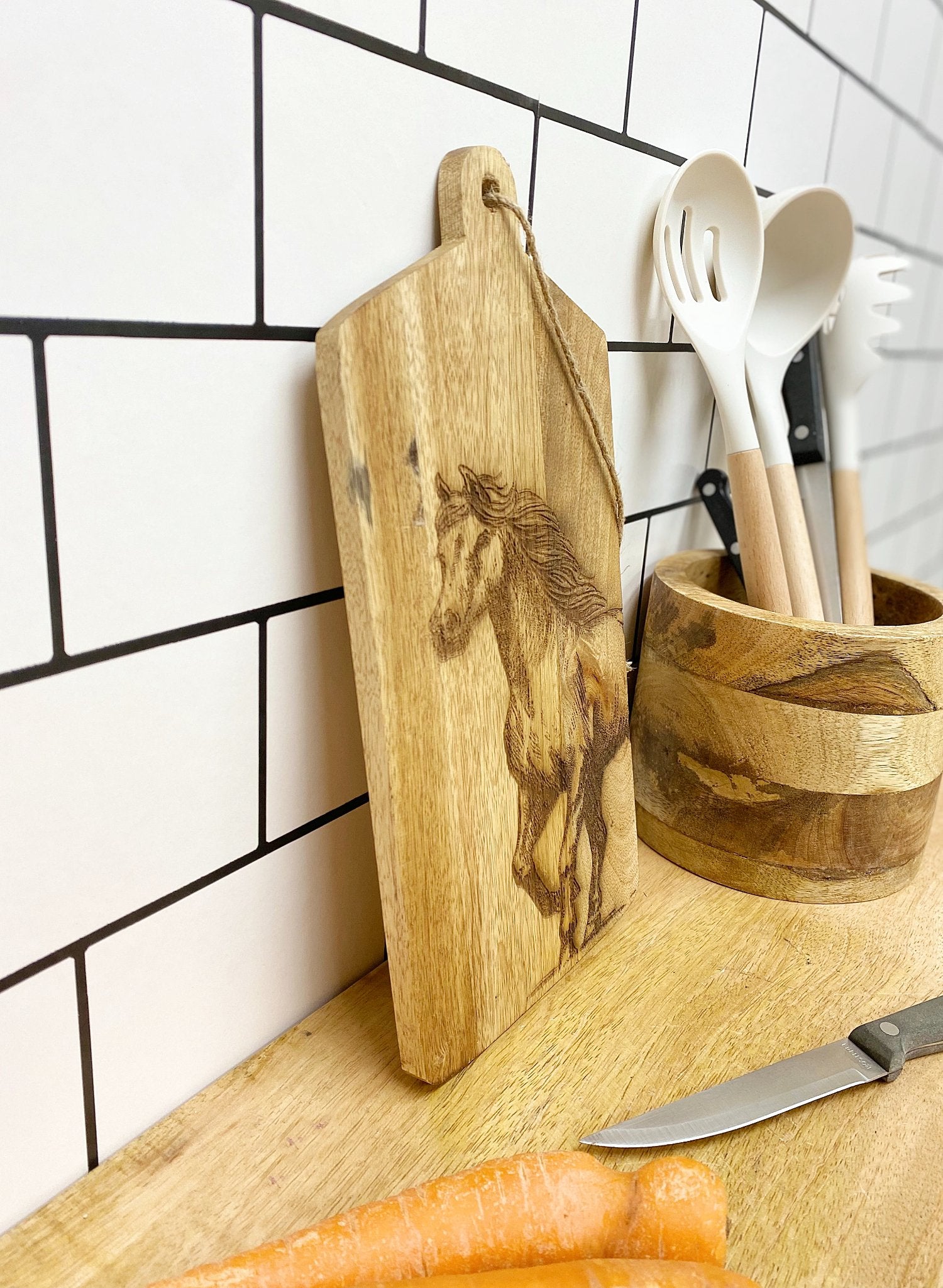 The Horse Engraved Wooden Cheese Board rests against a white tiled wall, beside a wooden utensil holder filled with spoons and spatulas. A knife and two carrots are on the wooden countertop.