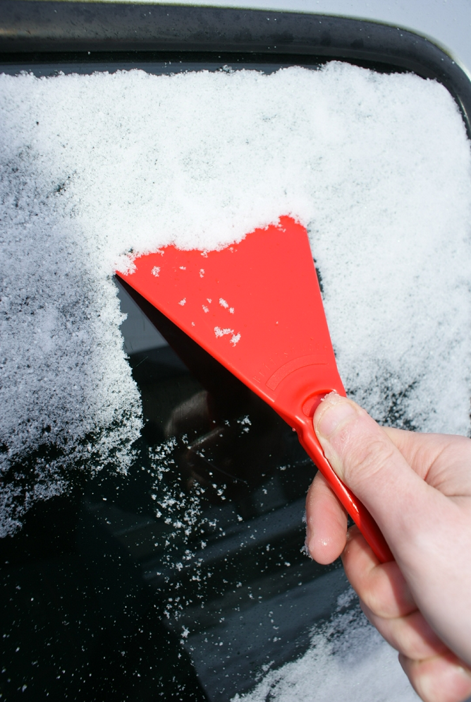 A hand uses the Essential Car Windscreen Ice Scraper, crafted from durable materials, to remove snow from the car window. The window is partially cleared, revealing the glass beneath surrounded by a layer of frost and ice.