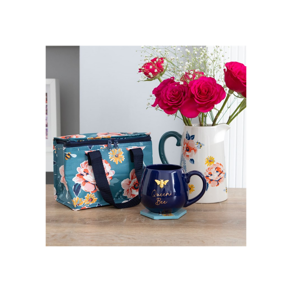 A Bee-utiful Floral Lunch Bag featuring blue fabric with pink and white floral patterns made from recycled plastic, a "Queen Bee" navy blue mug adorned with a bee illustration, and a white pitcher vase holding pink roses and baby's breath are arranged on a wooden table. The background includes a gray wall and part of a white doorframe.