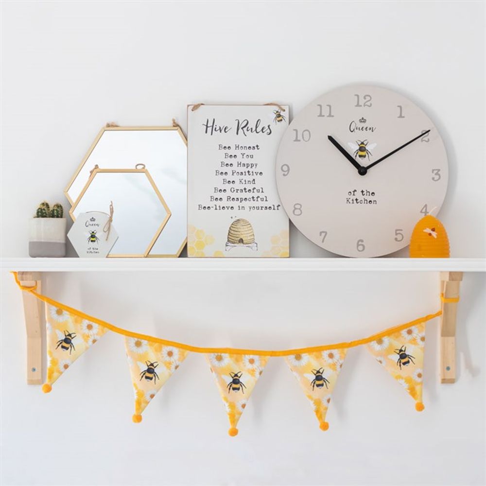 A white shelf with bee-themed home decor: a large geometric mirror, framed "Hive Rules," a bee-patterned clock, small potted plants, a candle, and a triangular bunting with bee designs hanging below.