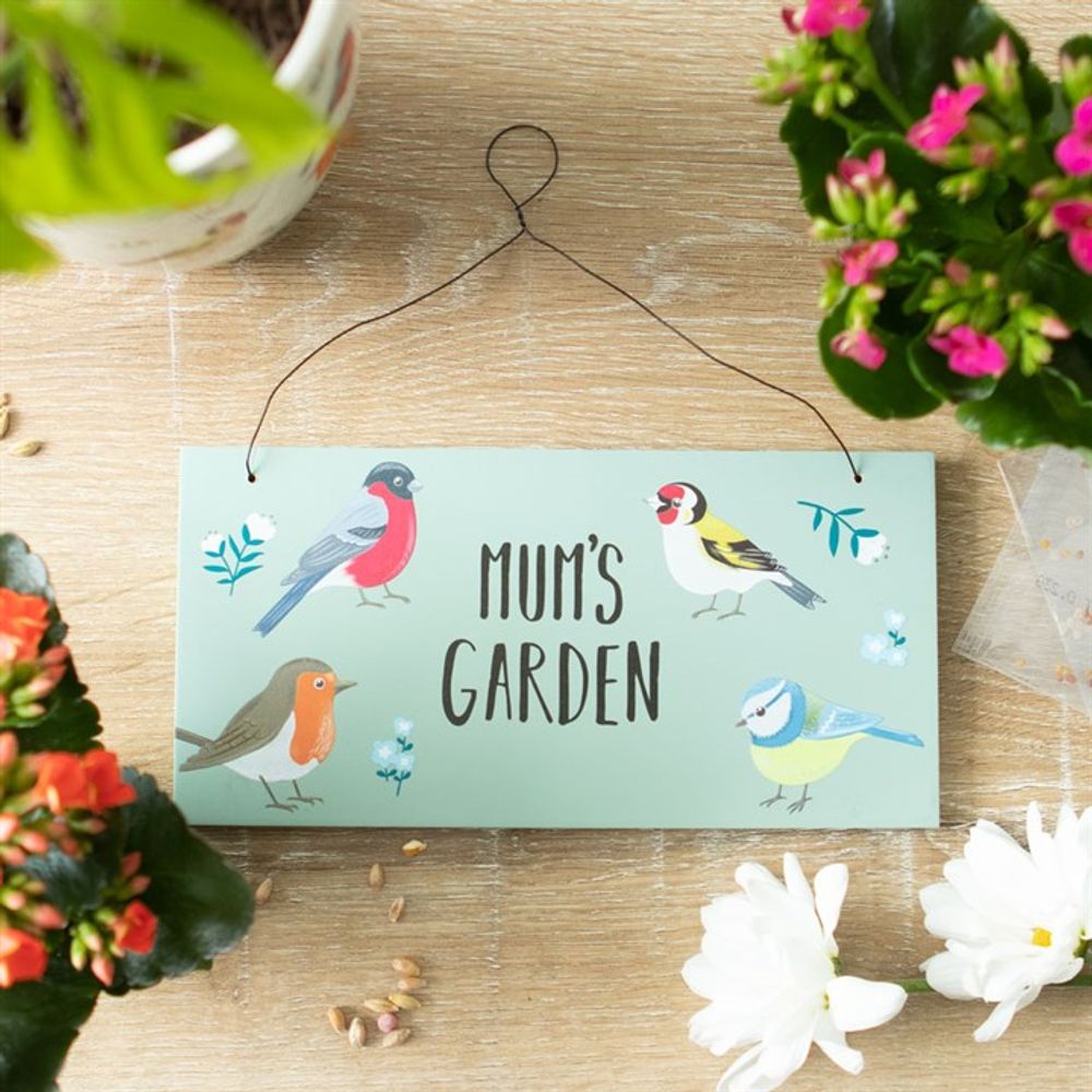 A "Mum's Garden British Garden Birds Sign," adorned with charming garden bird illustrations and wire hanger, rests on wood. Vibrant potted flowers with pink and white blooms frame the sign, while scattered seeds complete the whimsical scene.