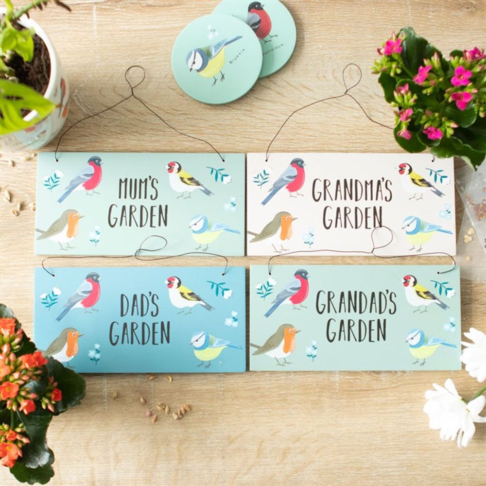 A wooden table displays four garden signs reading "Mum's Garden," "Dad's Garden," "Grandma's Garden," and "Grandad's Garden." Each sign features vibrant bird illustrations, with potted flowers adding colorful accents. The first sign is named Mum's Garden British Garden Birds Sign.