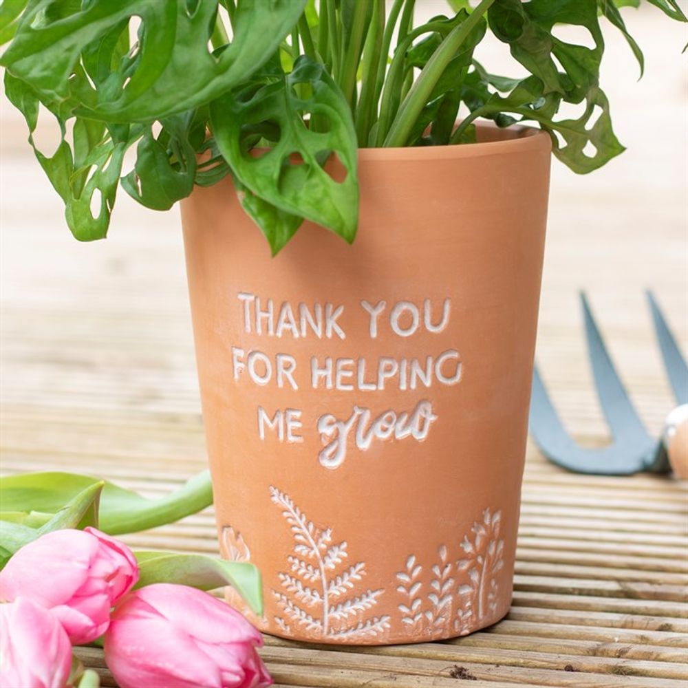 The Thank You For Helping Me Grow Terracotta Plant Pot has white engraved text and comes with a leafy plant inside. Pink tulips and a gardening tool sit nearby on the wooden surface.