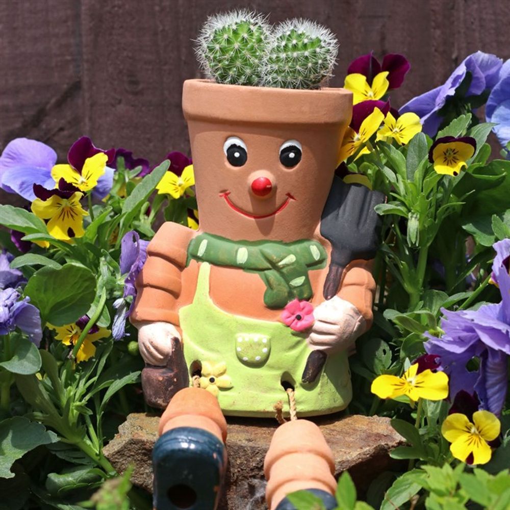 The Terracotta Pot Man Planter, with a whimsical face and green overalls, features two small cacti as hair and vibrant purple and yellow flowers, making it a charming addition perfect for brightening any garden.